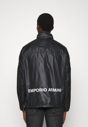 Osoba nosząca czarną, błyszczącą kurtkę z białymi literami "EMPORIO ARMANI" na dolnej części pleców, stojąca tyłem.