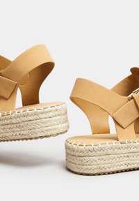 Sandali espadrilles marroni con una fascia larga, dettaglio a fiocco e piattaforma in giute intrecciato. Parte superiore in pelle liscia e suola testurizzata offrono un contrasto.