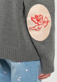 Gray knitted sweater featuring a rounded blush patch with red embroidered cherub, set against light blue floral-patterned jeans.