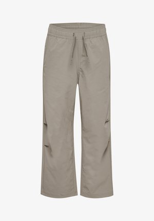 Barn iført grå elastiske bukser, hvide og sorte sneakers og en blå langærmet trøje, står mod en ensfarvet baggrund.