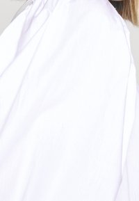 Camicia bianca in tessuto leggero e testurizzato. Presenta un colletto arricciato e maniche drappeggiate per un volume maggiore.