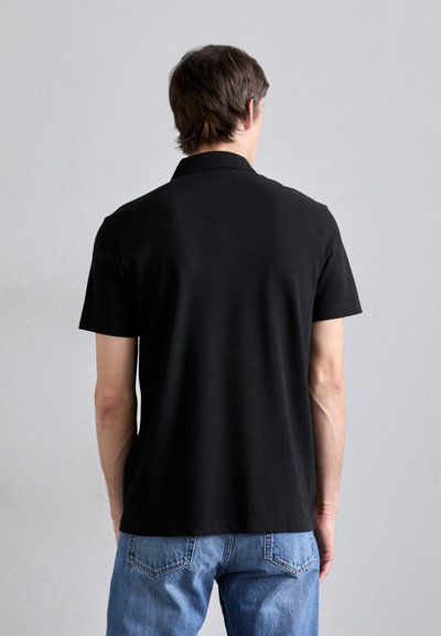 Polo shirt noir à manches courtes avec un col plat, texture lisse et design simple. Porté avec un jean bleu clair.