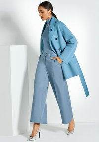 Vrouw in een lichtblauwe jas over de schouders, bijpassende blauwe coltrui en op maat gemaakte broek, geassisteerd met zilveren hakken en oorbellen met hoops.