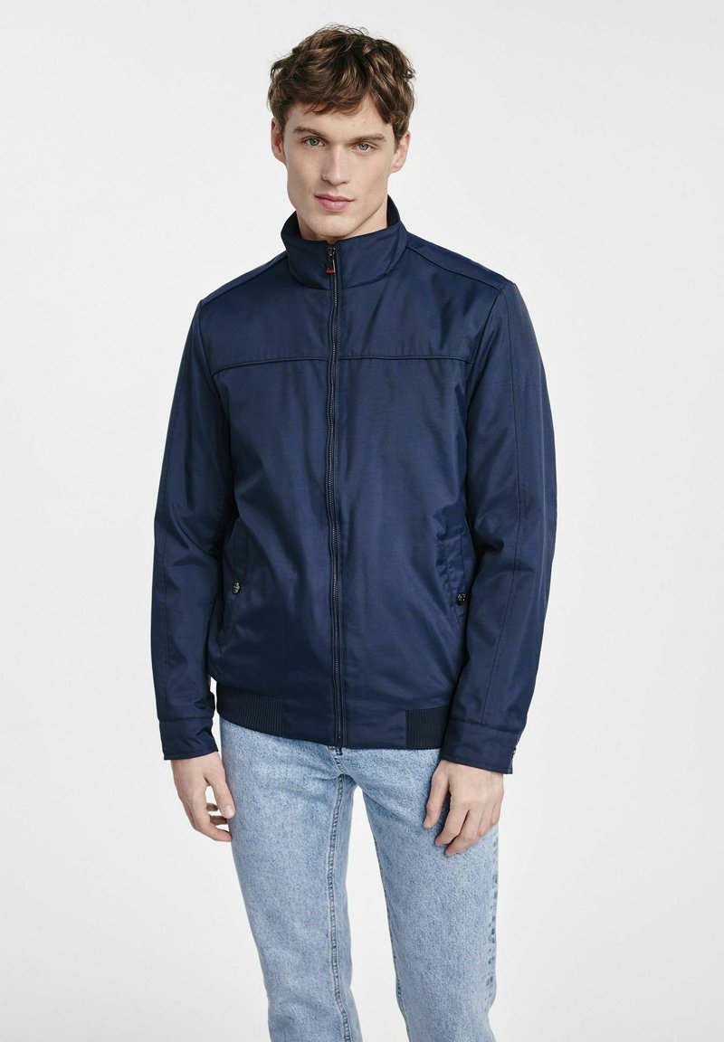 Granatowa kurtka zapinana na zamek z kołnierzem typu stojącego, bocznymi kieszeniami i ściągaczami. Gładka tekstura, lekki materiał i proste detale przeszyć.