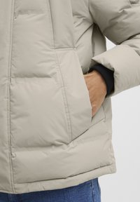 Beige puffer jacket featuring a textured finish, side pockets, and ribbed cuffs; visible stitching adds detail to the design.