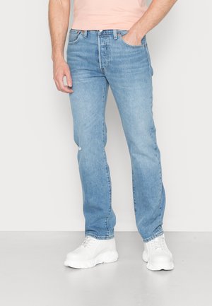 Homme portant un jean droit bleu clair et des baskets blanches épaisses, debout avec une main dans la poche sur un fond uni.