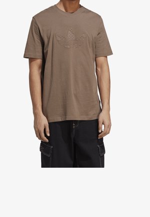 Homme portant un t-shirt marron à manches courtes avec un logo en relief à trois feuilles et un pantalon cargo noir avec des surpiqûres blanches.
