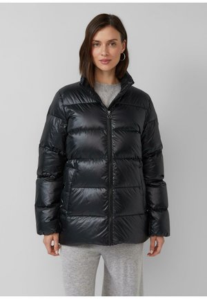 Femme portant une doudoune noire brillante avec fermeture éclair à l'avant et poches latérales, associée à un pantalon en tricot gris clair, se tenant contre un fond uni.