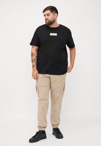 Czarny bawełniany t-shirt z prostokątną metką z logo, zestawiony z jasnobeżowymi spodniami cargo z bocznymi kieszeniami i elastycznymi mankietami. Czarne sneakersy.