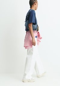 Junge Person mit geflochtenem Haar trägt weite weiße Hosen, ein marineblaues T-Shirt, ein pinkes Hemd um die Taille gebunden, eine denim Schultertasche undSneaker.