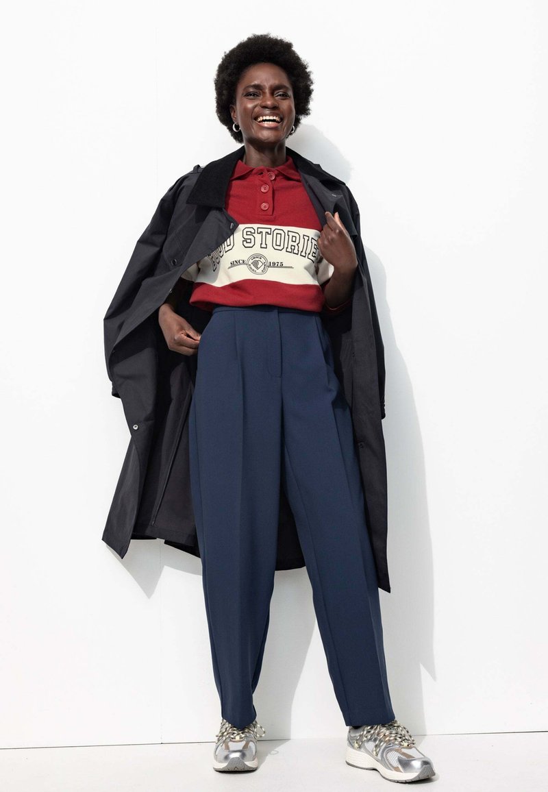 Sweatshirt rayé rouge et blanc avec logo, manteau trench noir, pantalon bleu marine taille haute, et baskets argentées avec lacets à motifs.