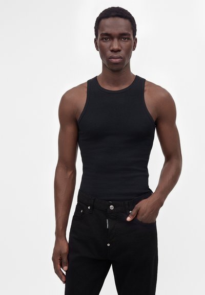 Homme portant un haut noir moulant sans manches et un pantalon noir, debout avec une main dans la poche sur un fond clair uni.