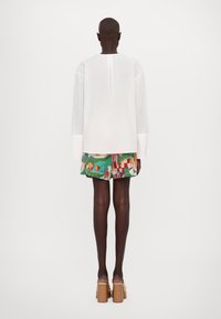 Haut blanc à manches longues côtelé avec un dos ouvert, associé à un short à motifs multicolores avec un fond vert et différentes formes. Sandales à talons carrés marron.