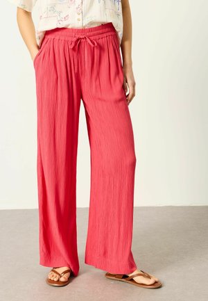 Personne portant un pantalon large rose vif avec une taille à cordon de serrage et des sandales marron, debout sur un sol gris devant un mur clair.