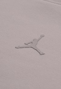 Ύφασμα ανοιχτού μωβ με υφή επιφάνειας που φέρει ένα γκρι κεντημένο λογότυπο Jumpman σε στάση άλματος, τοποθετημένο προς το κέντρο.