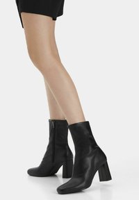Schwarze Leder-Stiefeletten mit hohem, quadratischem Absatz, ausgestattet mit einem seitlichen Reißverschluss und glatter Textur. Das Design ist schlank und körperbetont.