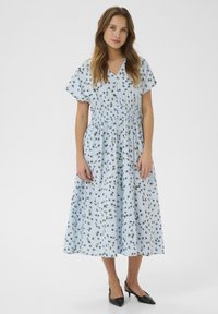 Ljusblå blommig klänning med korta ärmar, rynkad midja och en svepande kjol. Svarta spetsiga klackskor kompletterar outfiten.