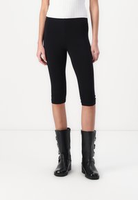 Juodos capri leggingai, pagaminti iš tampraus audinio, su lygiu paviršiumi ir prigludusia forma. Dėvimi su juodais odiniais batais iki kelių.
