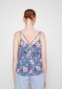 Camiseta sin mangas de patrón floral en tela azul, con tirantes finos y espalda en forma de V. El diseño incluye gráficos de flores rosas y moradas.