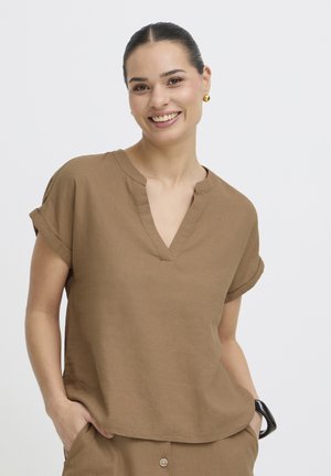 Žena s tamnom kosom u smeđoj bluzi s kratkim rukavima i V-izrezom te odgovarajućim hlačama, smiješi se s rukama u džepovima na jednobojnoj pozadini.