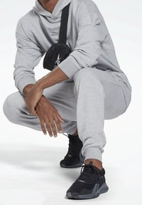 Person hockt, trägt hellgraue Jogginghose und Kapuzenpullover, schwarze Sneakers und eine schwarze Reebok-Umhängetasche.