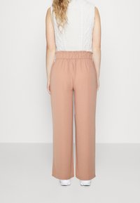 Žena stojící zády, oblečená v bílé bezrůžkové pletené topu, volných růžových kalhotách s vysokým pasem a bílých teniskách na jednolitém pozadí.