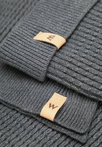 Szara dzianina prążkowana z dwoma skórzanymi etykietami, jedna oznaczona stylizowaną literą "A", a druga literą "W". Miękka tekstura i warstwowy design.