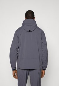 Giacca tecnica grigia con cappuccio, caratterizzata da una texture liscia e raffinata, polsini regolabili e collo alto per proteggere dal vento.