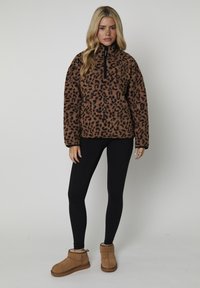 Donna che indossa un pullover in pile leopardato marrone, leggings neri e stivaletti beige, in piedi contro uno sfondo neutro.