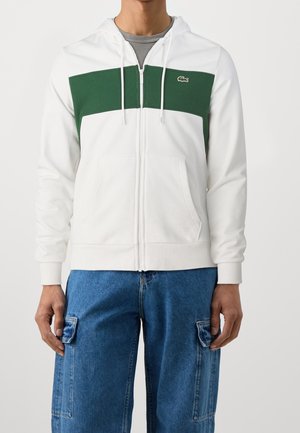 Person iført en hvid hættetrøje med lynlås med et vandret mørkegrønt panel og jeans med cargolommer, stående mod en ensfarvet baggrund.