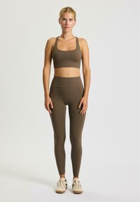 Top corto sportivo marrone e leggings a vita alta, realizzati in materiale elasticizzato, design aderente, texture liscia, abbinati a scarpe da ginnastica chiare.