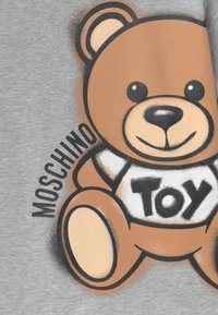 Šedá tkanina obsahuje grafiku medvěda v hnědé a béžové barvě, který nosí bílou košili s černým nápisem "TOY". Design zahrnuje zaoblené tvary a jemné linie.