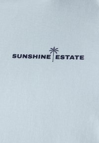 Svetle modrá tkanina se hladkým povrchem obsahuje text "SUNSHINE ESTATE" v tmavě námořnické barvě, doplněný jednoduchou grafikou palmy.