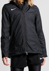 Femme portant une veste zippée noire Nike et un short noir Nike avec des leggings noirs sur un fond gris uni.