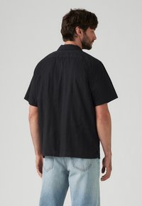 Zwart kortemouwen shirt met een subtiele textuurpatroon. Heeft een klassieke kraag en een rechte zoom, gecombineerd met lichtblauwe jeans.