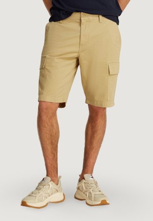 Homme portant un short cargo beige et des baskets crèmes chunky, se tenant contre un fond clair uni.