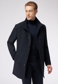 Cappotto di lana navy con ampio colletto, chiusura a bottone monopetto, due tasche laterali e una finitura testurizzata. Abbinato a un dolcevita coordinato.