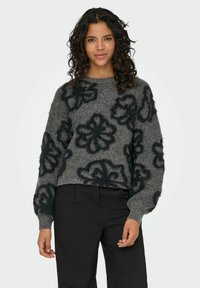 Frau mit lockigem Haar, die einen grauen Pullover mit gro�ßen schwarzen Blumenmustern und schwarze Hosen trägt, steht vor einem schlichten Hintergrund.
