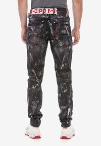 Schwarze Jeans aus Denim mit einem gespritzten Farbmuster in Weiß und Rot. Verfügt über einen weißen Taillenbund mit Logo. Sichtbare Gesäßtaschen und Nähte.