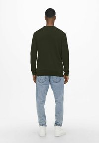 Camisa de manga larga verde oscura con cuello redondo, combinada con jeans azul claro y zapatillas blancas. Enfócate en el corte relajado y la textura de la tela.