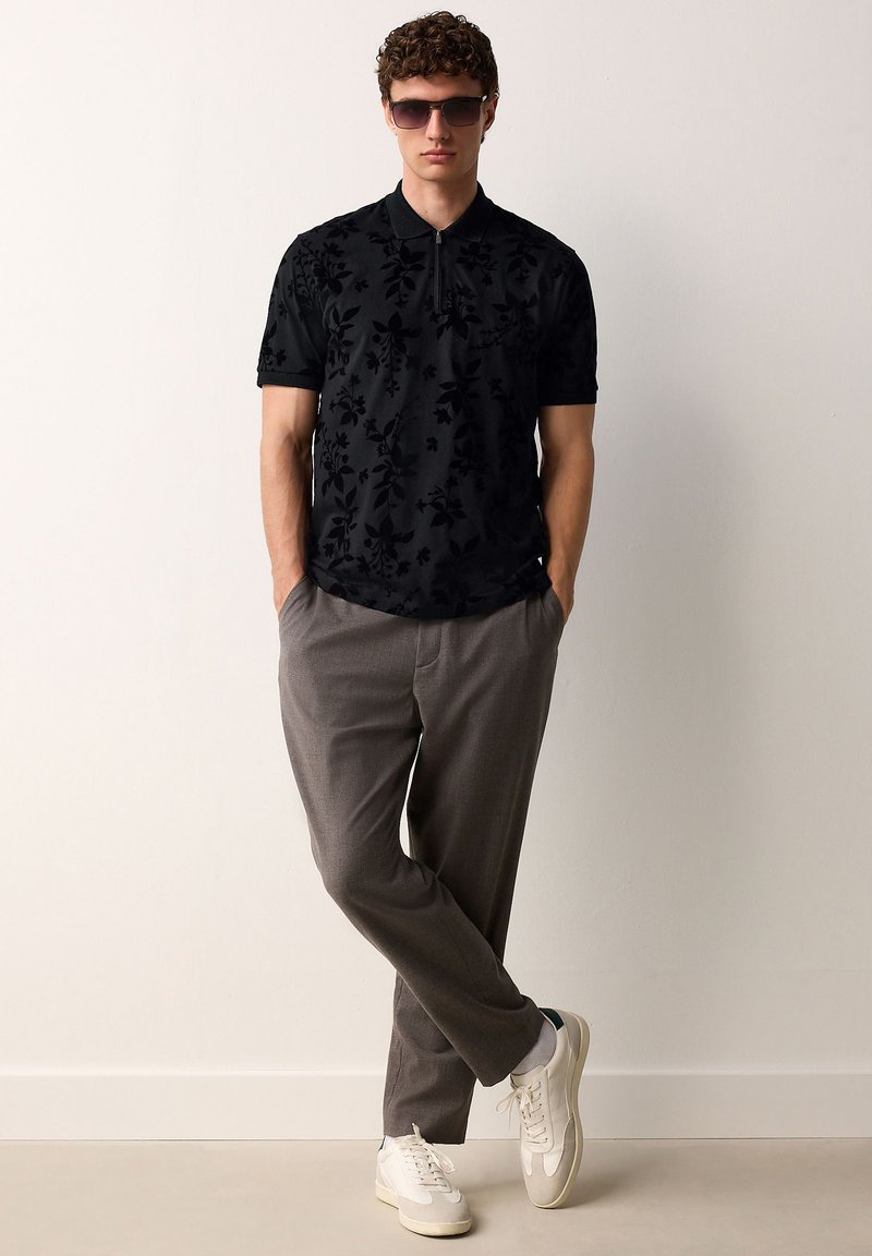 Férfi sötét színű, virágmintás fekete galléros pólóban, szürke nadrágban, fehér tornacipőben és napszemüvegben, zsebébe dugott kézzel áll.