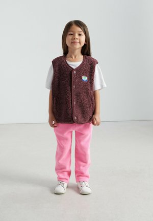 Pruun fliisvest nööpidega, kantud valge T-särke peal, kombineeritud roosade fliispükstega ja valgete vabaajajalatsitega. Tasapinnaline, tekstuuriga pind.