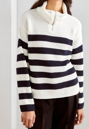 Woman wearing a white and black horizontal striped knit sweater with a half zipper and high collar, paired with black pants.