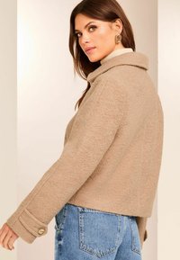 Giacca cropped beige con finitura testurizzata, dotata di colletto e polsini con bottoni, abbinata a jeans blu a vita alta.