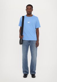 Blå bomull T-skjorte med kvadratisk MK-logo, kombinert med lyseblå rette jeans og svarte sko, holder en svart skulderveske.