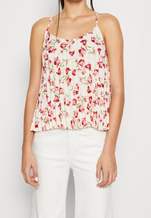 Kvinde iført en ærmeløs, plisseret hvid top med røde og pink blomsterprint og hvide bukser, med håret sat op.