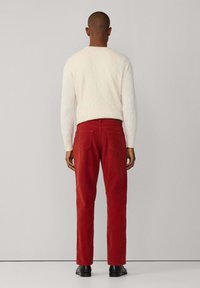 Pantalones de algodón rojos y un suéter de punto trenzado color crema, vistos desde atrás, con zapatos negros. Textura suave, diseño ajustado y detalles mínimos.