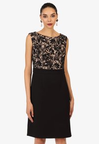 Robe noire avec un corsage en dentelle à motifs floraux et une jupe unie et ajustée. Design sans manches et coupe au genou. Encolure élégante.