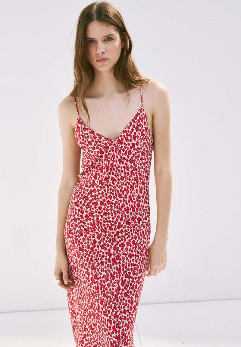Jeune femme portant une robe nuisette fleurie rouge et blanche sans manches, debout devant un fond clair uni.