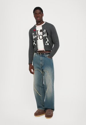 Jonge man in een grijze cardigan met hondenpatroon, witte graphic tee, blauwe spijkerbroek met hartvormige naadontwerpen en bruine geborduurde schoenen.